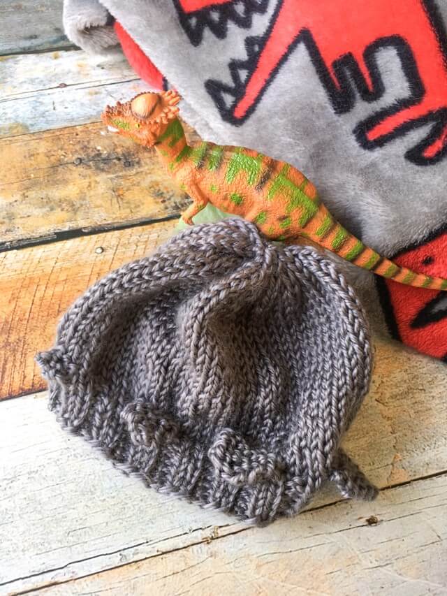 A gray knitted baby hat styled after a pachycephalosaurus lays against a t rex blanket and wooden background with a toy pachycephalosaurus next to it.