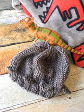 Load image into Gallery viewer, A gray knitted baby hat styled after a pachycephalosaurus lays against a t rex blanket and wooden background with a toy pachycephalosaurus next to it.
