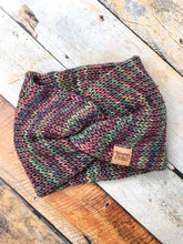 Load image into Gallery viewer, A knit headband with knot in the front shown against a wooden background.  The headband is a blend of rainbow colors, mostly pink and green.
