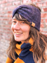 Load image into Gallery viewer, Knitted headband with knotted front shown on a model against a brick background.  The headband is a blend of dark blues and purples.
