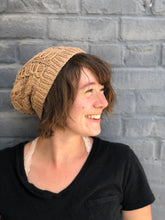 Load image into Gallery viewer, The Fossil Beanie is shown here in worsted weight, knitted with cotton for a light summer slouch.  It is shown in light brown on a model against a blue background.
