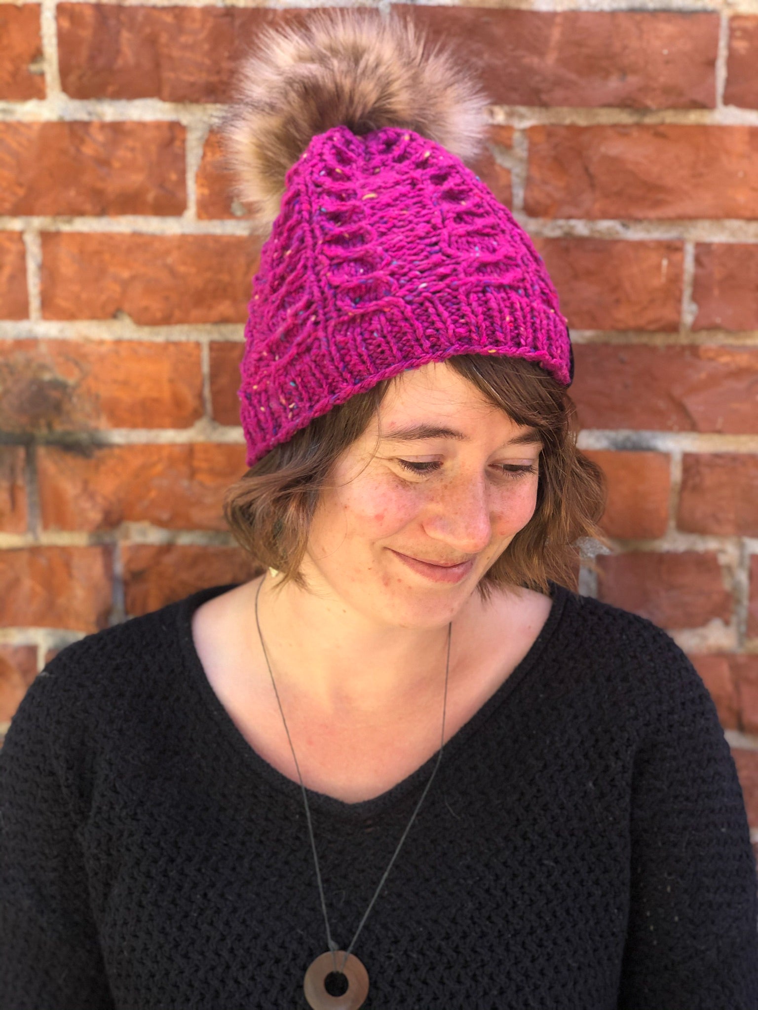 The Fossil Beanie is shown here in bulky yarn in magenta tweed with a brown pom.  It is shown on a model.