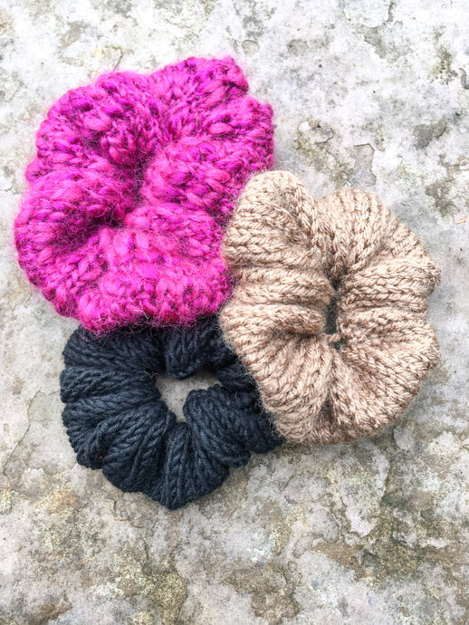 Three knit scrunchies are shown in a flat lay.  They are magenta, light brown, and black.