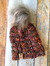 Load image into Gallery viewer, Witch Hazel Beanie in brown with gray pom. It is shown here in a flat lay against a wooden background.
