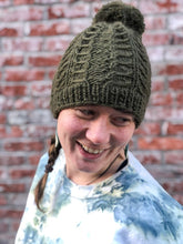 Load image into Gallery viewer, Fossil Beanie in olive with matching yarn pom.  It is shown here on a model.
