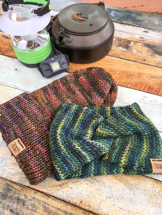 A folded brim slouchy beanie in flat lay with a knotted head wrap.  The hat has pink and green tones.  The wrap has yellow and blue tones.  In the background are camping supplies:  a kettle, a lantern, and a headlamp.