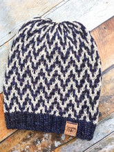 Load image into Gallery viewer, The Find Your Way Beanie has a chevron stripe pattern in two colors.  It is shown here in a flat lay in white and navy.
